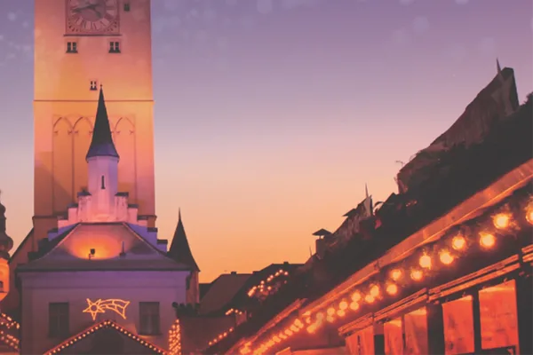 Die Dächer eines beleuchteten Weihnachtsmarkts und ein beleuchteter Kirchturm in der Abenddämmerung.