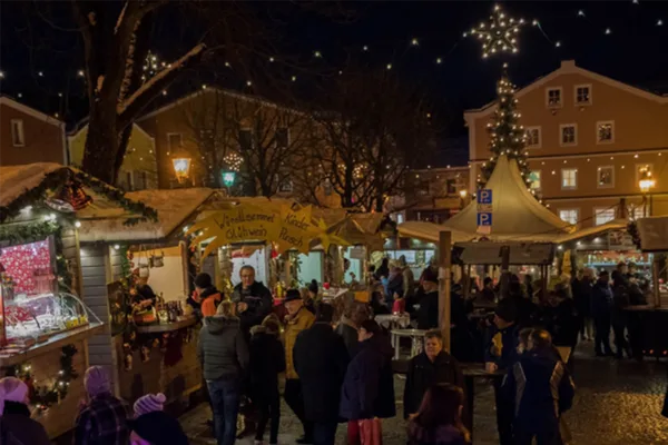 Ein hell erleuchteter Weihnachtsmarkt auf einem Stadtplatz mit einzelnen Ständen und vielen Besuchern im Dunkeln.