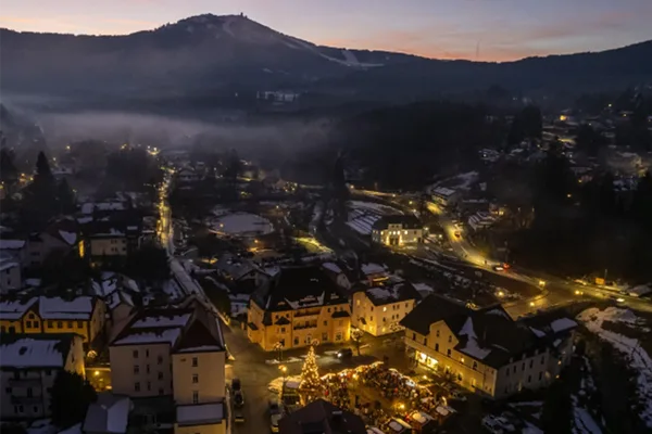 Blick von oben auf einen Stadtplatz mit beleuchteten Weihnachtsmarktständen. Im Hintergrund ist noch der Sonnenuntergang hinter den Berggipfeln zu erkennen.