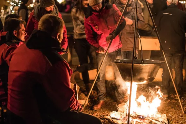 Menschen sitzen um ein Lagerfeuer, über dem ein Topf mit einem Dreibein aufgehängt wurde.