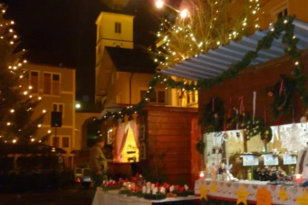 Ein Weihnachtsbaum mit Lichterkette neben Weihnachtsmarktbuden, die mit Tannengirlanden und Lichterketten geschmückt sind.