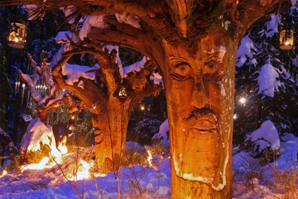 Mehrere beleuchtete Baumstämme, in die große Gesichter geschnitzt wurden, in einer verschneiten Waldlandschaft.