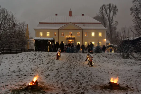 Ein mit kleinen Lagerfeuern beleuchteter Weg über eine verschneite Wiese zu einem großen Gebäude, vor dem ein kleiner Weihnachtsmarkt aufgebaut ist.