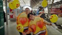 Auf diesem Bild sehen Sie zwei Promoter:innen, die große Emoji-Kostüme tragen und Luftballons halten.