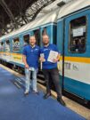 Auf diesem Bild sehen Sie zwei Mitarbeiter der Länderbahn, die Werbemateralien in der Hand halten.
