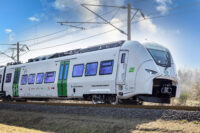 Sie sehen auf diesem Bild den Siemens Mireo Zug von vorn während einer Fahrt. Im Hintergrund sind nicht belaubte Bäume.