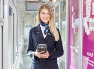 Eine lächelnde Zugbegleiterin steht mit einem Verkaufsterminal in der Hand im Gang eines Reisezugwagens.