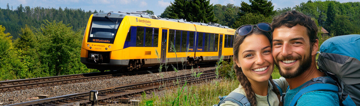 Header Bahn und wandern mit der oberpfalzbahn Ein Mann und eine Frau in Wanderkleidung stehen auf einer Wiese und lächeln in die Kamera. Im Hintergrund sind Schienen zu sehen auf denen ein gelber Zug der Oberpfalzbahn fährt.