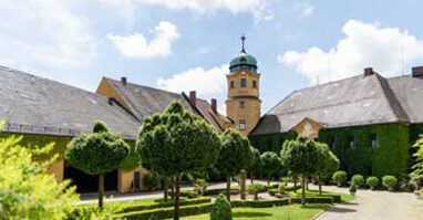 Schloss Reuth Reuther Kulturrunde Das Bild zeigt das Schloss Reuthe im Sommer bei Sonnenschein. Im Vordergrund stehen Bäume in einer Parkanlage.