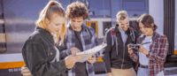 Eine Gruppe von Personen schaut in eine Wanderkarte und in eine Kamera am Bahnhof.