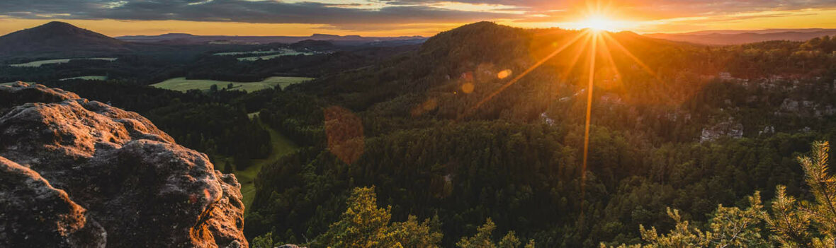 Das Bild zeigt eine malerische Landschaft mit sanften Hügeln und dichten Wäldern. Die Sonne geht gerade auf oder unter und taucht die Szene in ein warmes, goldenes Licht. Im Vordergrund sind einige Felsvorsprünge zu sehen, und der Himmel ist teilweise bewölkt mit orangefarbenen und gelben Tönen vom Sonnenlicht.