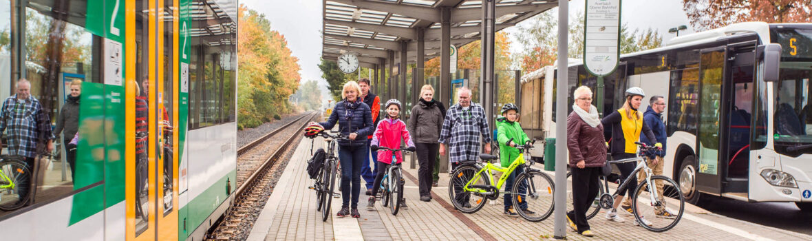 Das Bild zeigt eine Gruppe von Menschen, die auf einem Bahnsteig stehen. Einige Personen haben Fahrräder dabei. Links im Bild steht ein Zug der vogtlandbahn, rechts ein Bus. Der Bahnsteig hat eine Dachkonstruktion mit Glasplatten, und im Hintergrund sind Bäume mit Herbstlaub zu sehen.
