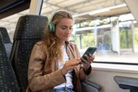 Das Bild zeigt eine Person, die auf einem Sitzplatz im Zug sitzt, Kopfhörer trägt und ein Smartphone benutzt. Die Person trägt eine braune Lederjacke und blaue Jeans. Im Hintergrund ist der Bahnsteig des Bahnhofs durch das Fenster zu sehen.