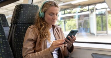 Pendlerin im Zug Das Bild zeigt eine Person, die auf einem Sitzplatz im Zug sitzt, Kopfhörer trägt und ein Smartphone benutzt. Die Person trägt eine braune Lederjacke und blaue Jeans. Im Hintergrund ist der Bahnsteig des Bahnhofs durch das Fenster zu sehen.