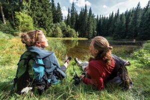 Klingenthal Höhensteig Das Bild zeigt zwei Personen, die auf dem Gras nahe einem kleinen See oder Teich im Hüttenbachtal sitzen, umgeben von einem dichten Wald. Die Person links trägt eine blaue Jacke und hat einen grün-schwarzen Rucksack, während die Person rechts ein rotes Shirt trägt und einen grauen Rucksack hat. Beide blicken auf das Wasser, mit Bäumen im Hintergrund unter einem teilweise bewölkten Himmel.