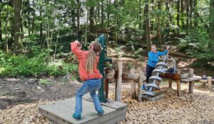Schoeneck Moosmaennleinweg Das Bild zeigt zwei Kinder, die in einem Outdoor-Spielplatzbereich spielen, umgeben von Bäumen und Laub. Ein Kind steht auf einer Holzplattform und bedient eine grüne Handpumpe, während das andere Kind mit einem spiralförmigen Wasserspiel interagiert. Der Boden ist mit Holzspänen bedeckt, und es gibt verschiedene Holzstrukturen und Elemente um sie herum.