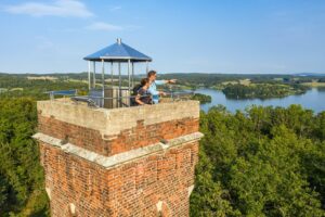 Elster und Triebtal Mosenturm Talsperre Das Bild zeigt einen Backstein-Aussichtsturm mit einem Metalldach und einem Geländer oben. Zwei Personen stehen auf der Aussichtsplattform und blicken auf eine malerische Landschaft mit dichten grünen Wäldern und einem großen Gewässer im Hintergrund. Der Himmel ist klar mit einigen verstreuten Wolken.