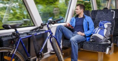 Reisender mit Fahrrad im Zug Das Bild zeigt einen Mann, der in einem Zug sitzt und aus dem Fenster schaut. Neben ihm steht ein Fahrrad, das sicher an einer Halterung befestigt ist. Er trägt eine blaue Jacke und Jeans, während sein Rucksack auf dem Sitz neben ihm liegt. Der Zug fährt durch eine grüne Landschaft.