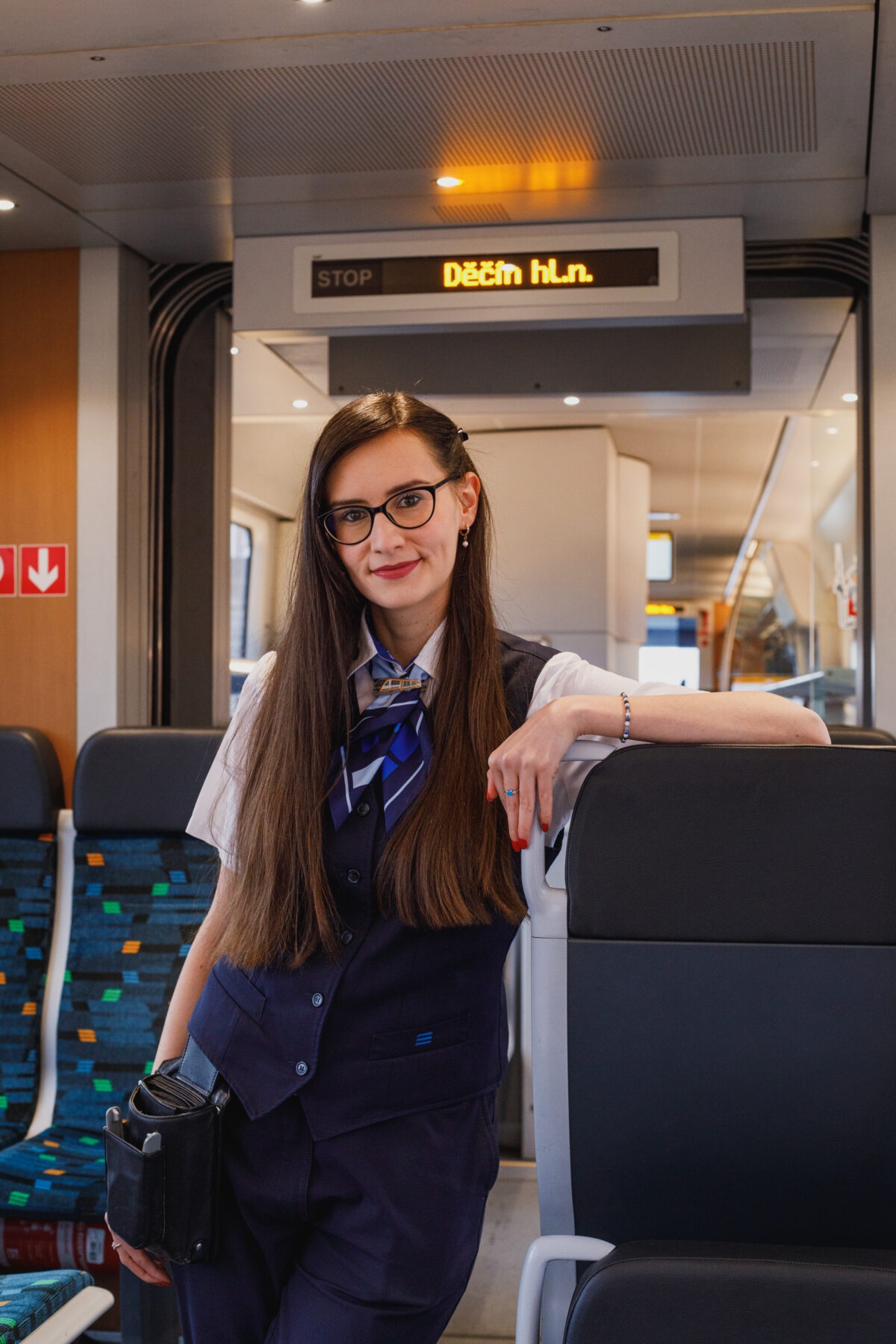 Na obrázku je slečna průvodčí. Stojí v uličce ve vlaku trilex, opírá se o sedadlo a usmívá se.