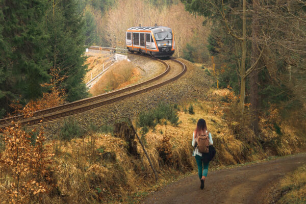Vlak trilex projíždí přírodou, vpravo po turistické cestě jde turistka.